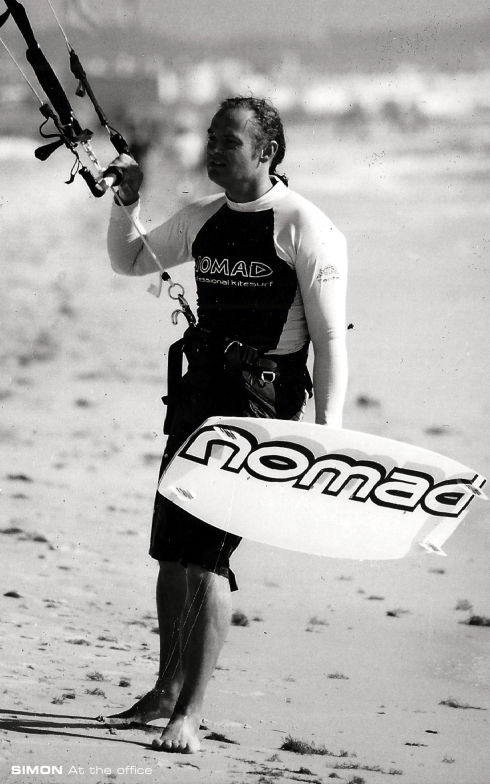 Simon carrying a Nomad board on the beach, captioned 'Simon at the office'
