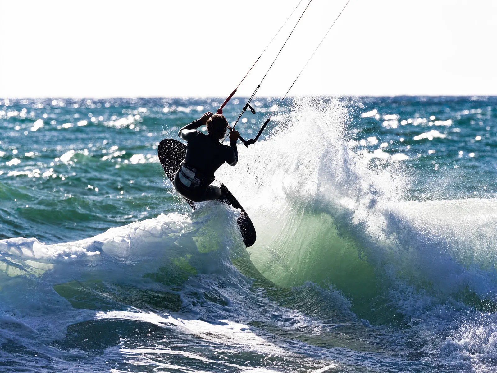 Backlit silhouette of a Nomad Wave Twin-Tip rider carving hard off a sunlit breaking wave, board rail dug in and spray exploding behind